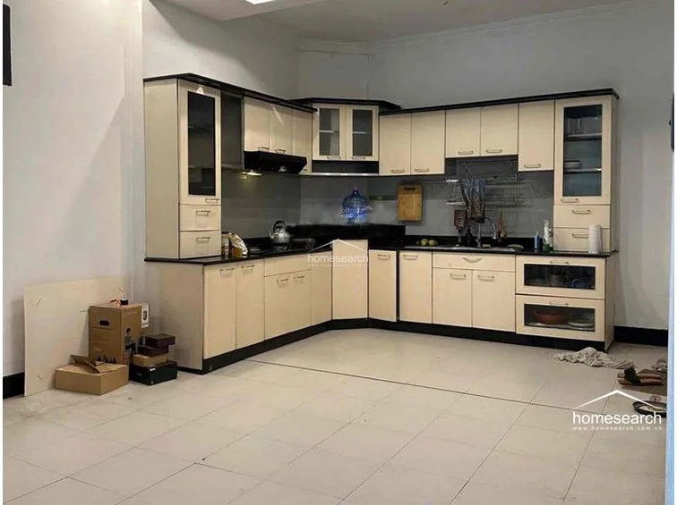 The kitchen features an L-shaped layout with light-toned cabinetry contrasted by dark countertops and trim, providing ample storage and prep space. The room is finished with neutral floor tiles and white walls, creating a clean, functional environment that appears in good condition.