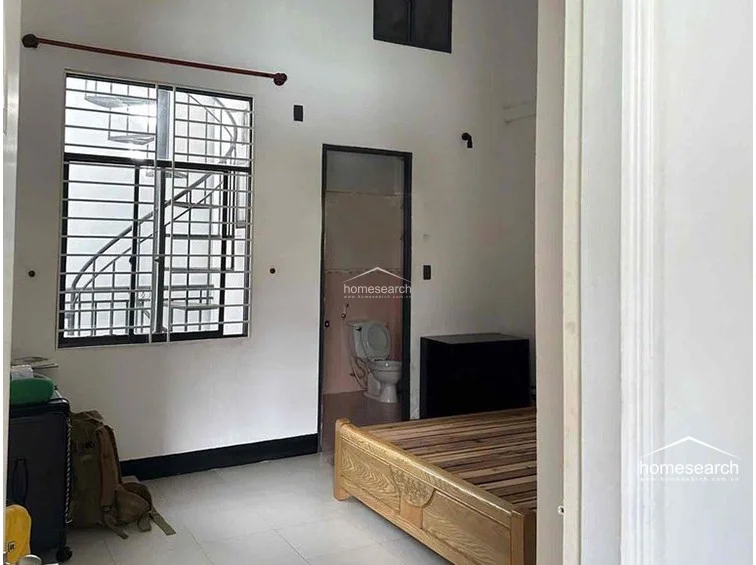 This bedroom includes an en-suite bathroom and features a large window with security bars that overlooks an external spiral staircase. The room is furnished with a wooden bed frame and is finished with simple white walls and light-colored floor tiles, reflecting a functional and straightforward design.