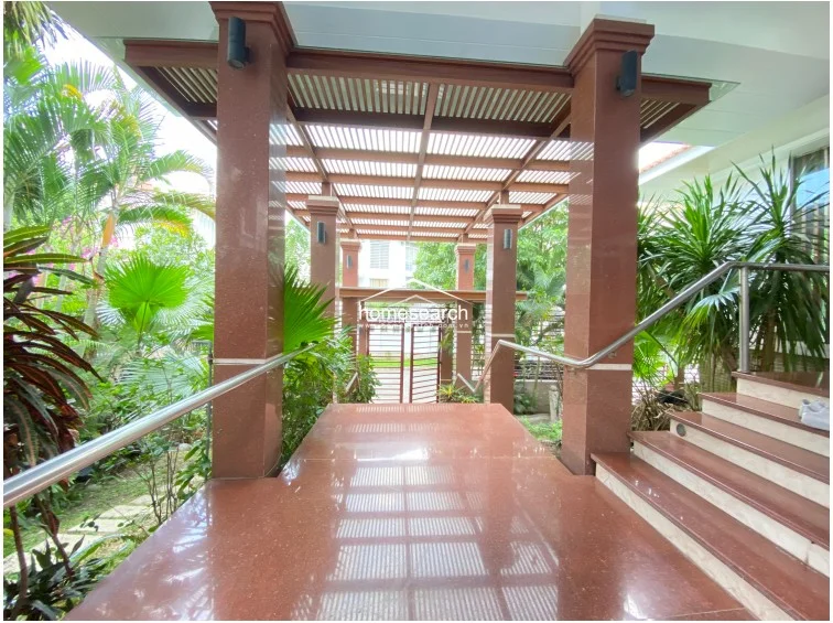 This image focuses on the grand entrance area, featuring a polished red granite floor and matching structural columns that create a sophisticated, high-end aesthetic. A slatted wooden pergola overhead provides filtered sunlight, while a stainless steel railing ensures safety along the staircase leading to the interior.