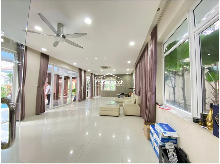 This spacious, open-concept living area features large floor-to-ceiling glass doors that provide excellent natural light and access to an outdoor terrace. The room is finished with high-gloss white tile flooring, recessed ceiling lighting, and a modern ceiling fan, creating a bright and airy atmosphere. A neutral color palette is complemented by mauve curtains and a large beige sectional sofa, offering a clean and well-maintained living space.