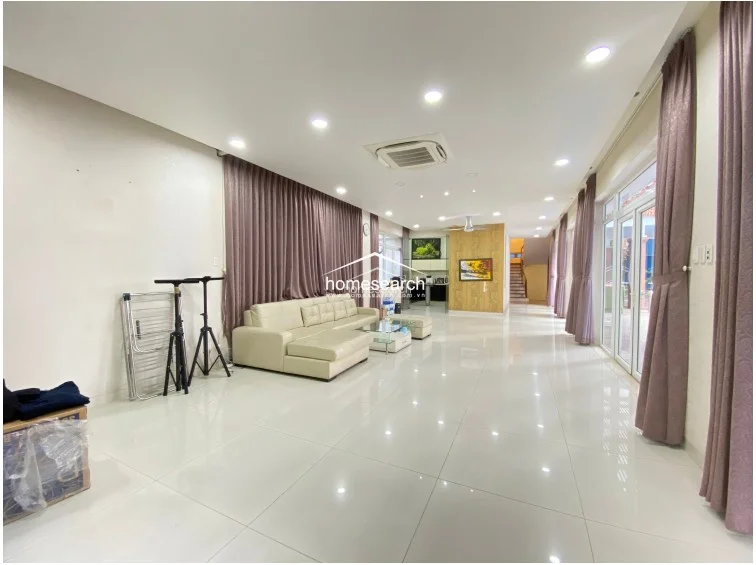 The living room maintains a spacious, airy layout with a neutral color palette and high-gloss tile floors that reflect the bright, natural light from the large windows. The space is furnished with a comfortable cream-colored sectional sofa and is equipped with a modern ceiling fan, offering a clean, well-maintained environment that connects easily to the adjacent pool area.