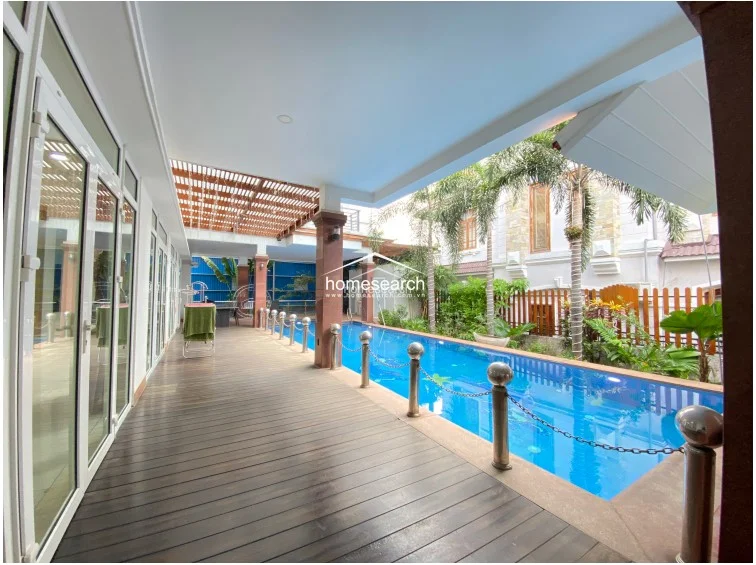 This outdoor space features a long, rectangular swimming pool bordered by a dark wood-plank deck and a covered patio area. The structure is supported by sturdy, reddish-brown columns and includes a decorative wooden slatted ceiling, providing a seamless transition between the indoor living areas and the lush, tropical garden landscape.