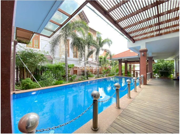 The pool area is well-maintained, featuring a clear blue swimming pool with a safety chain barrier and a secondary covered terrace with outdoor dining furniture. The space is surrounded by mature palm trees and greenery, creating a private, resort-like atmosphere that is ideal for relaxation or outdoor entertaining.