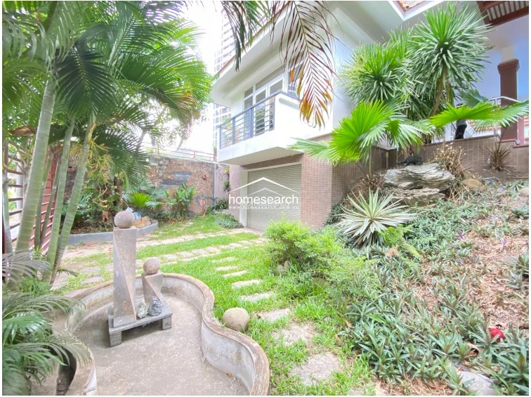 This exterior view showcases a lush, landscaped garden featuring a decorative stone water fountain and a stone-paved pathway. The property exterior is finished in neutral-toned tiling, and a roll-down security shutter covers what appears to be a ground-level garage or storage area.