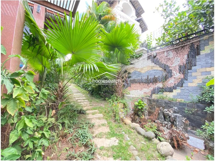 This shot captures a scenic garden path lined with vibrant tropical foliage, leading toward a decorative stone wall mural. The path is constructed with natural stone pavers set into the grass, creating a serene and well-maintained outdoor environment that integrates art with nature.