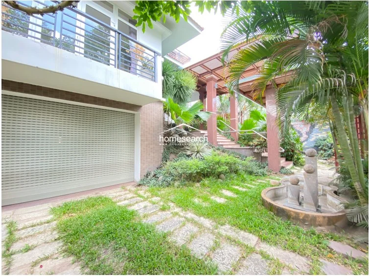 This angle highlights the transition from the garden to the upper levels, emphasizing the mature greenery and a stone-tiled staircase leading to the main entrance. The architecture incorporates a modern pergola structure supported by sturdy columns, providing a shaded walkway that connects different sections of the property.
